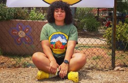 Raquel Gomez sits crossed legged on the ground with her eyes closed smiling at the camera. The sign behind her says Atabey Outdoors.