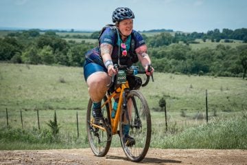 Marley Blonksy is riding a bike on a dirt path with a green field behind her.