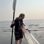 A man carries a lightweight kayak and paddle out of the water.