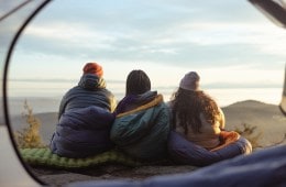 Three people sit in their sleeping bags looking out at a view in front of them.