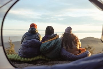 Three people sit in their sleeping bags looking out at a view in front of them.