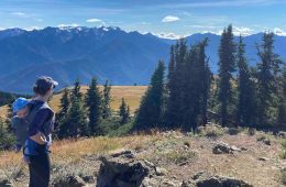 A hiker with a kid carrier on her back (holding her young son) looks off toward the mountains