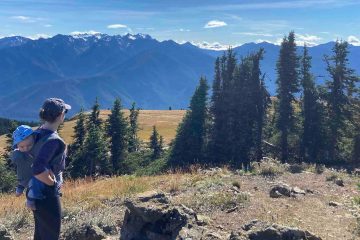 A hiker with a kid carrier on her back (holding her young son) looks off toward the mountains