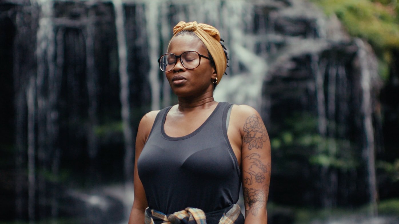 A Black woman wearing glasses, a headwrap and a tank top, standing in front of a waterfall.