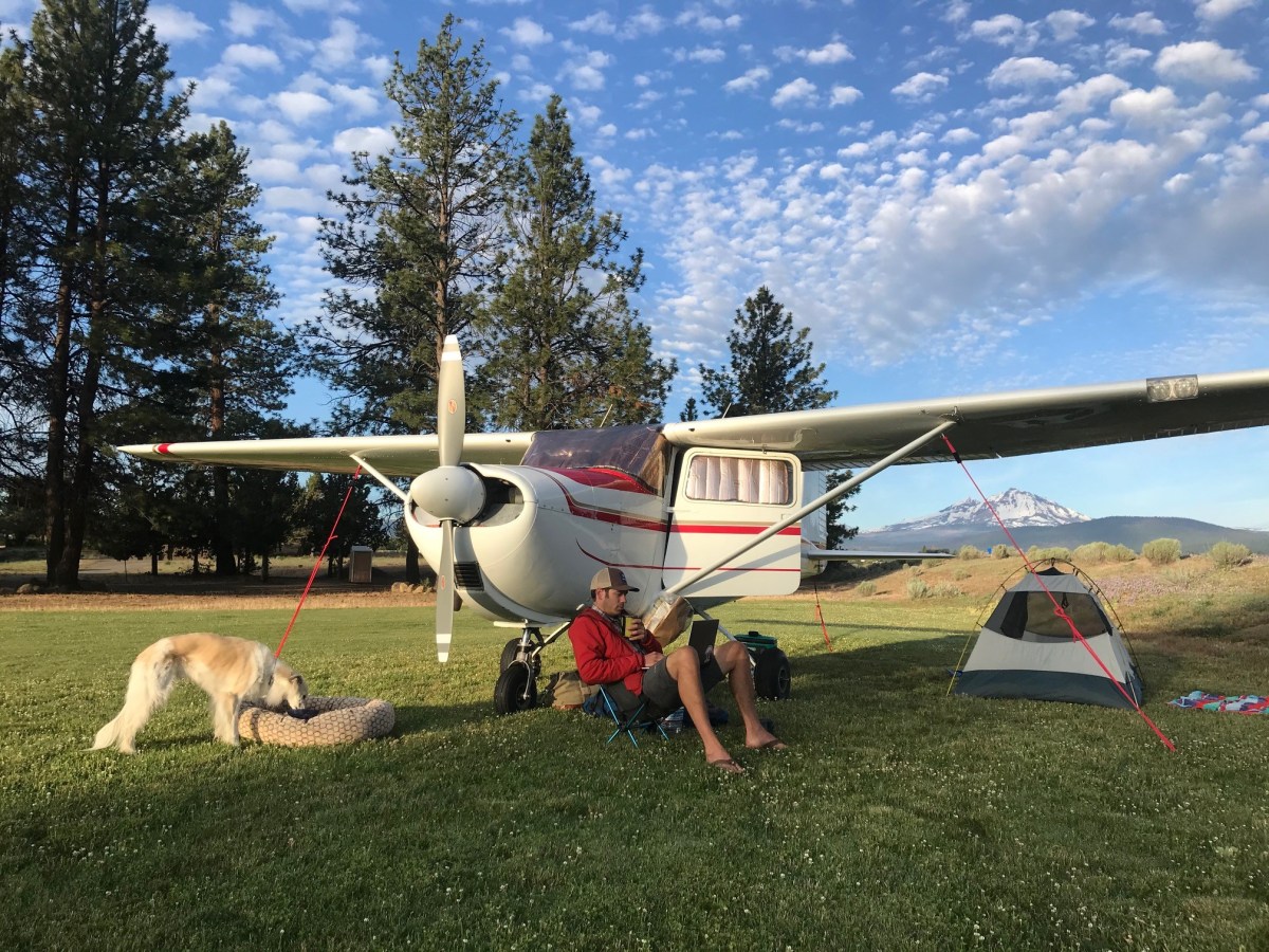 A man sits in a camp chair next to a small plane, his dog and a Half Dome Tent.