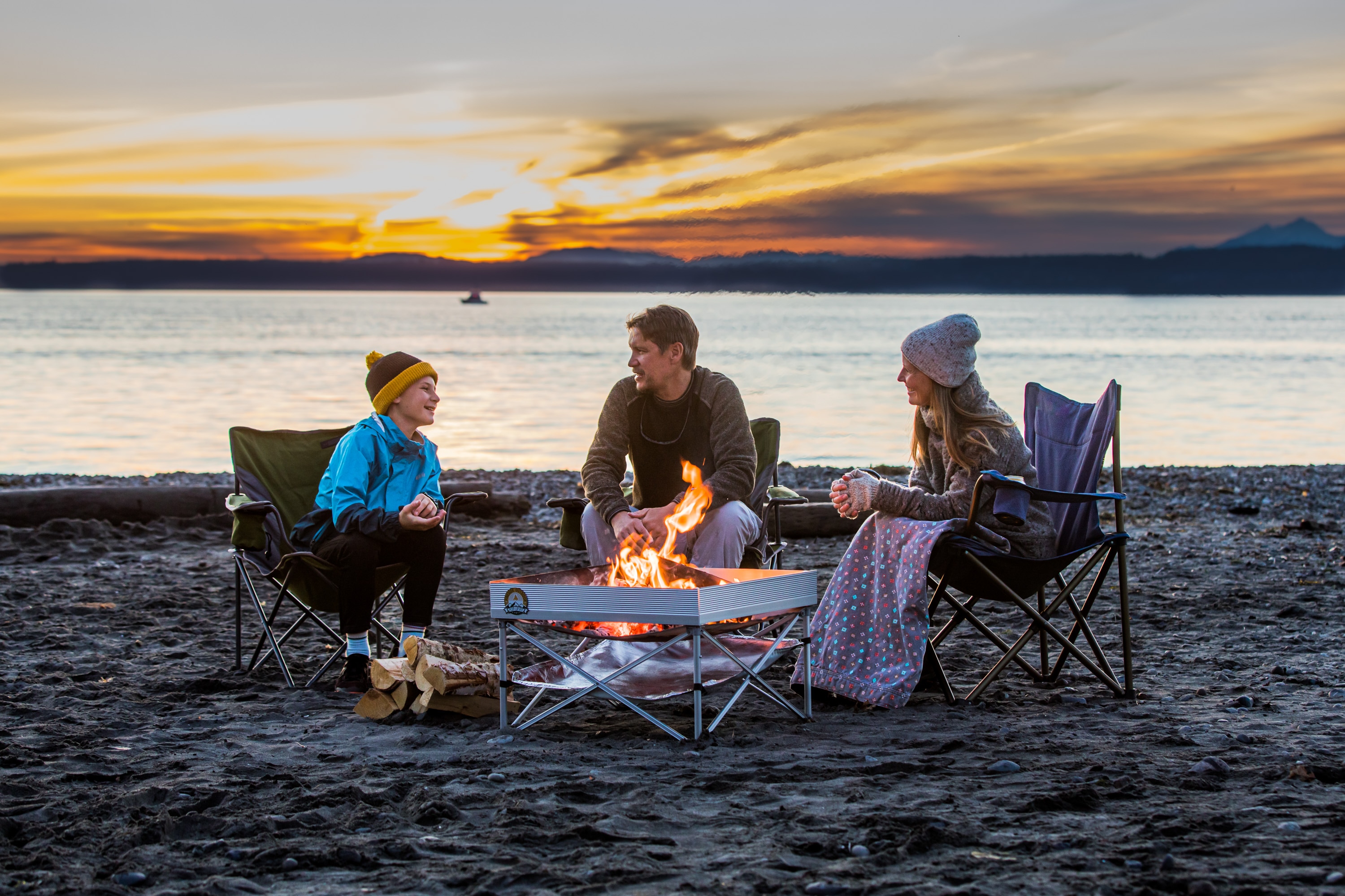 Three people enjoying time by the fire at the beach at sunset.