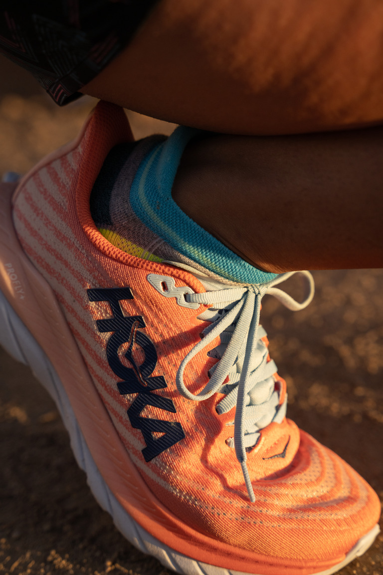 A person's lower leg, squatting, they're wearing the HOKA Mach 5 in peach and light blue