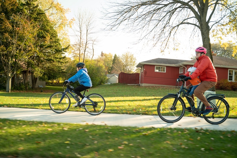 A child bikes in front of an adult on neighborhood sidewalk