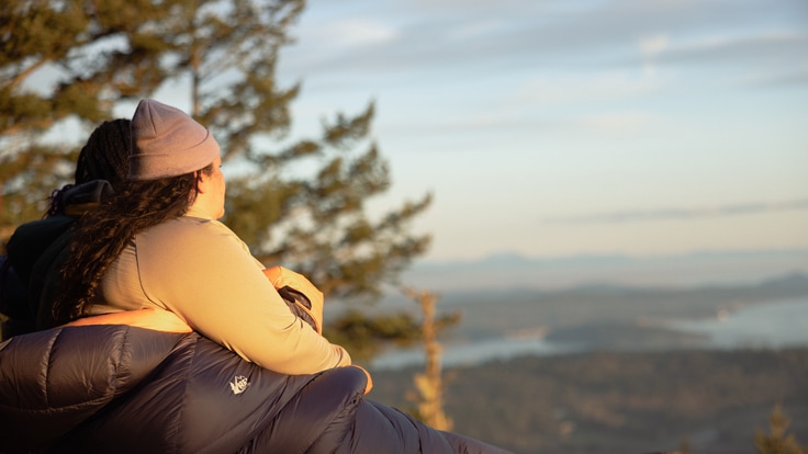 A person wrapped in a sleeping bag gazes out at the view from a mountain.