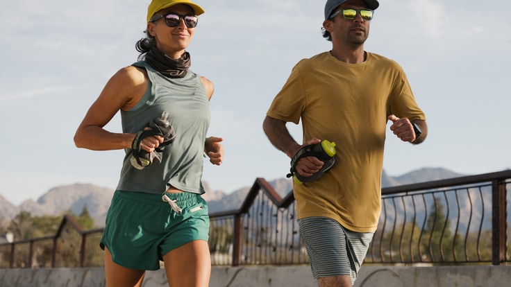 Two runners wearing sunglasses jog along a path. They are both wearing hats, summer shirts and shorts and carry sport water bottles.