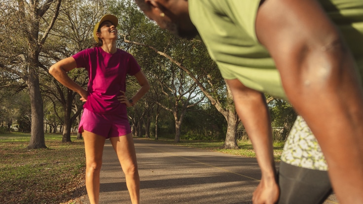 A person in a magenta running outfit and white running shoes smiles, looking up, with their hands on their hips, while another person in green and black running clothes rests with their hands on their knees in the foreground.