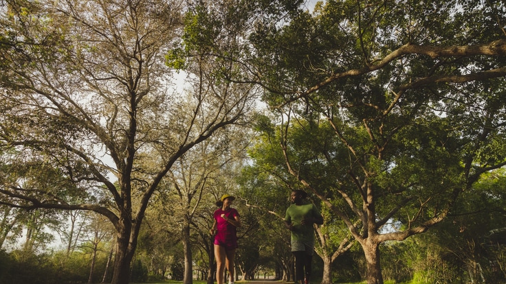 Two runners running towards the camera, one in a magenta running kit and the other in a green running kit, both wearing HOKA Clifton 9 road-running shoes.