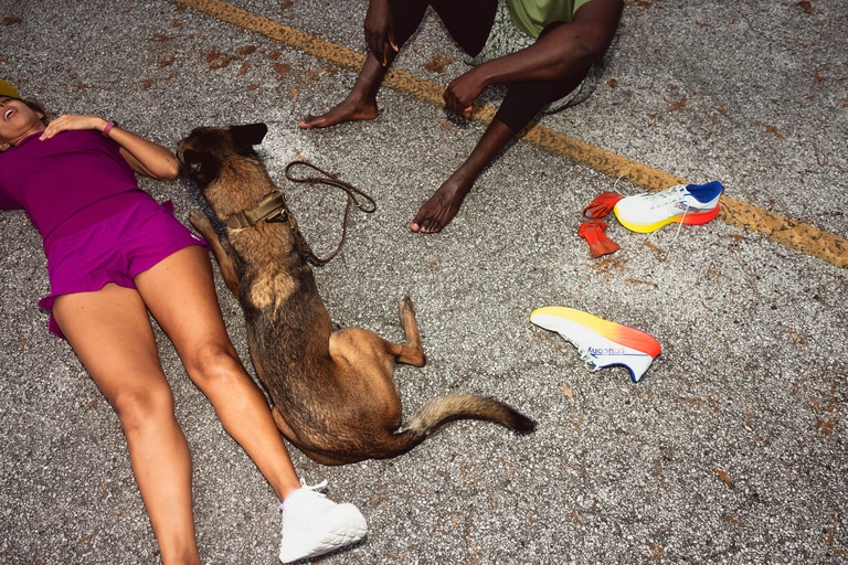 One person, in a magenta running outfit and white running shoes, lying down on the road next to a German Shepherd dog, who is also lying down. Another person sits, with only their legs and arms in view, next to them. The second person's running shoes and socks are off and splayed next to them.