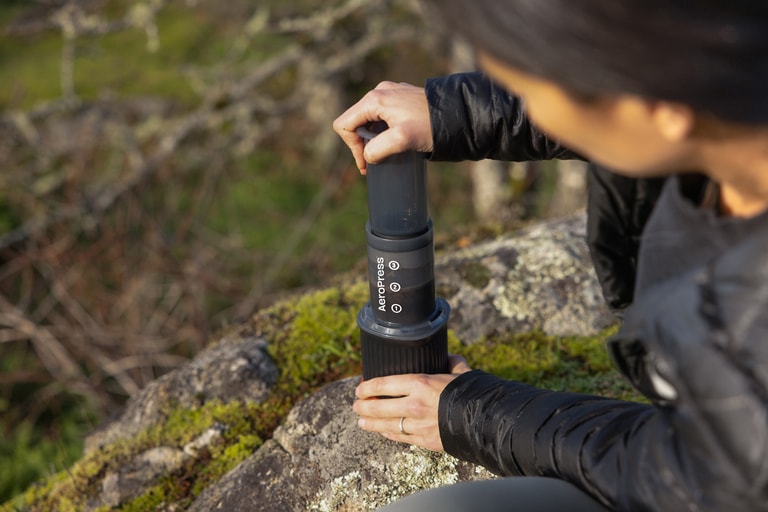 A person pressing down on the plunger of a coffee maker while it's resting on a rock