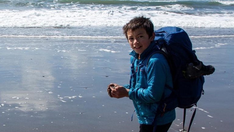 A boy wearing an insulated jacket carries a backpack along the beach