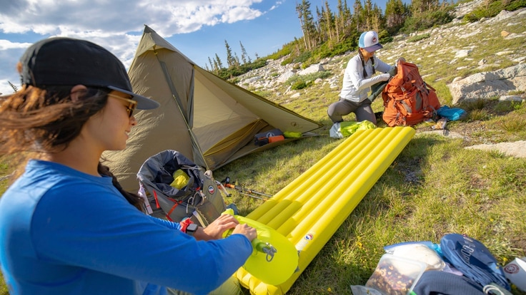 Two people at a campsite, with a tent, packs and a sleeping pad.