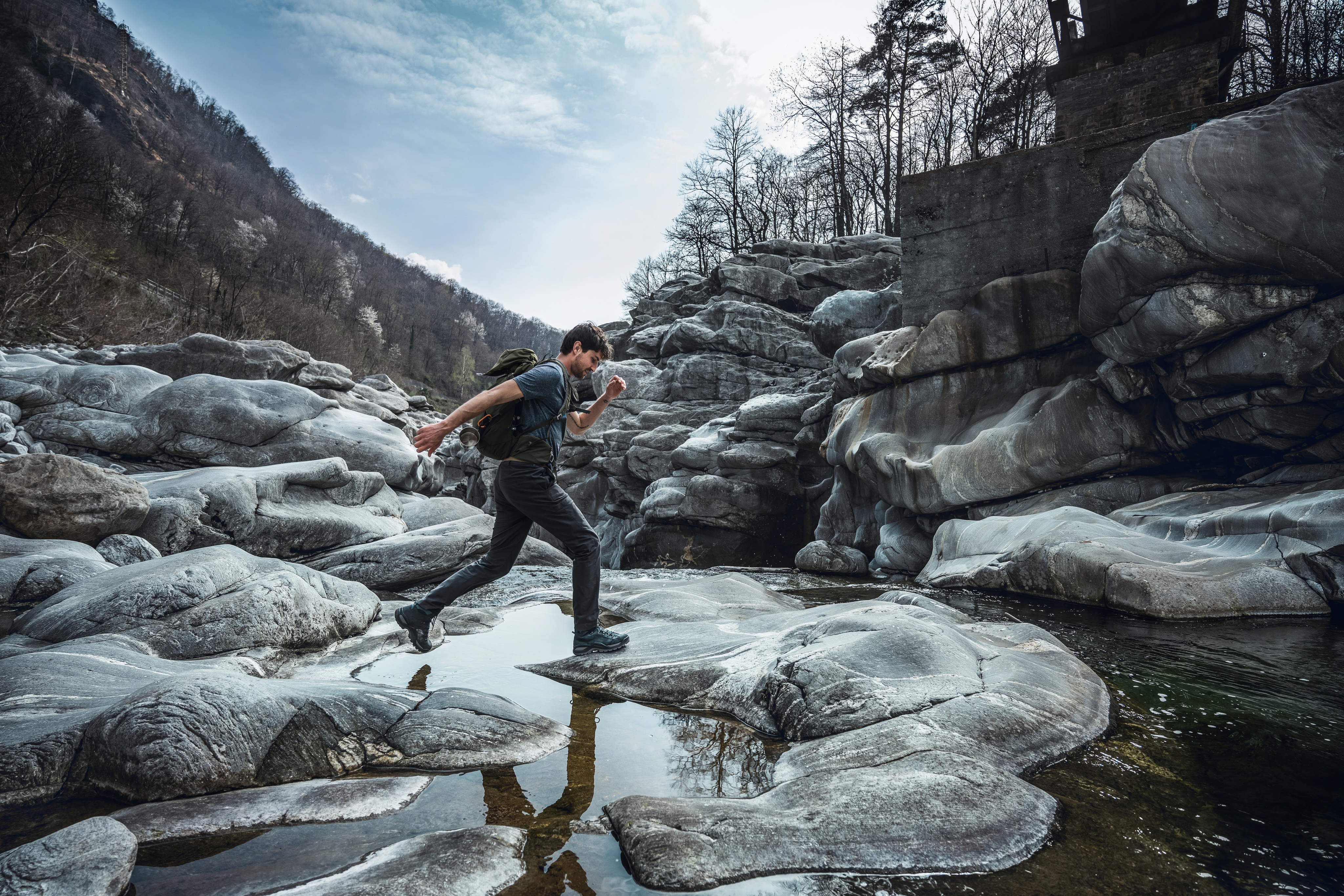 A person jumps across a shallow waterway on smooth gray rocks.