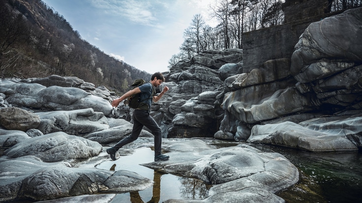 A person jumps across a shallow waterway on smooth gray rocks.