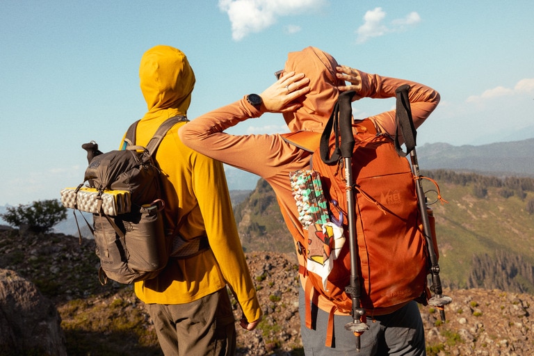 Two hikers stand with their backs to the camera, showing off their packs with sleeping pads and trekking poles strapped to them