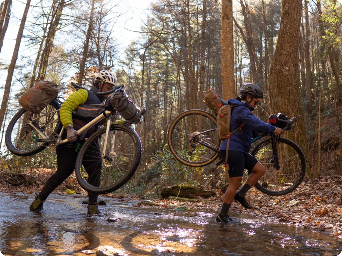 Two people carrying their bikes over a creek.