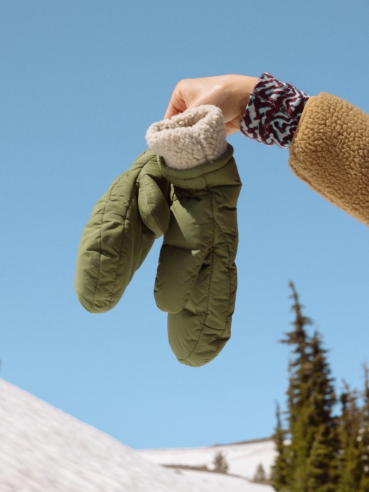 A person holds up winter gloves in front of a snowy landscape.