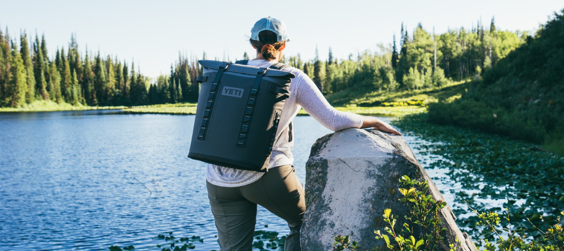 A person with a YETI backpack cooler stands by a rock, overlooking a peaceful lake.