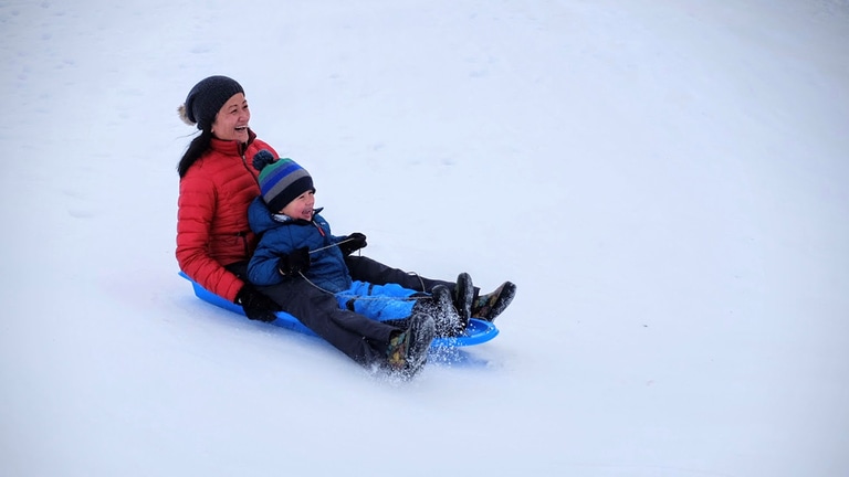 An adult and child smile while sledding down a snowy hill.