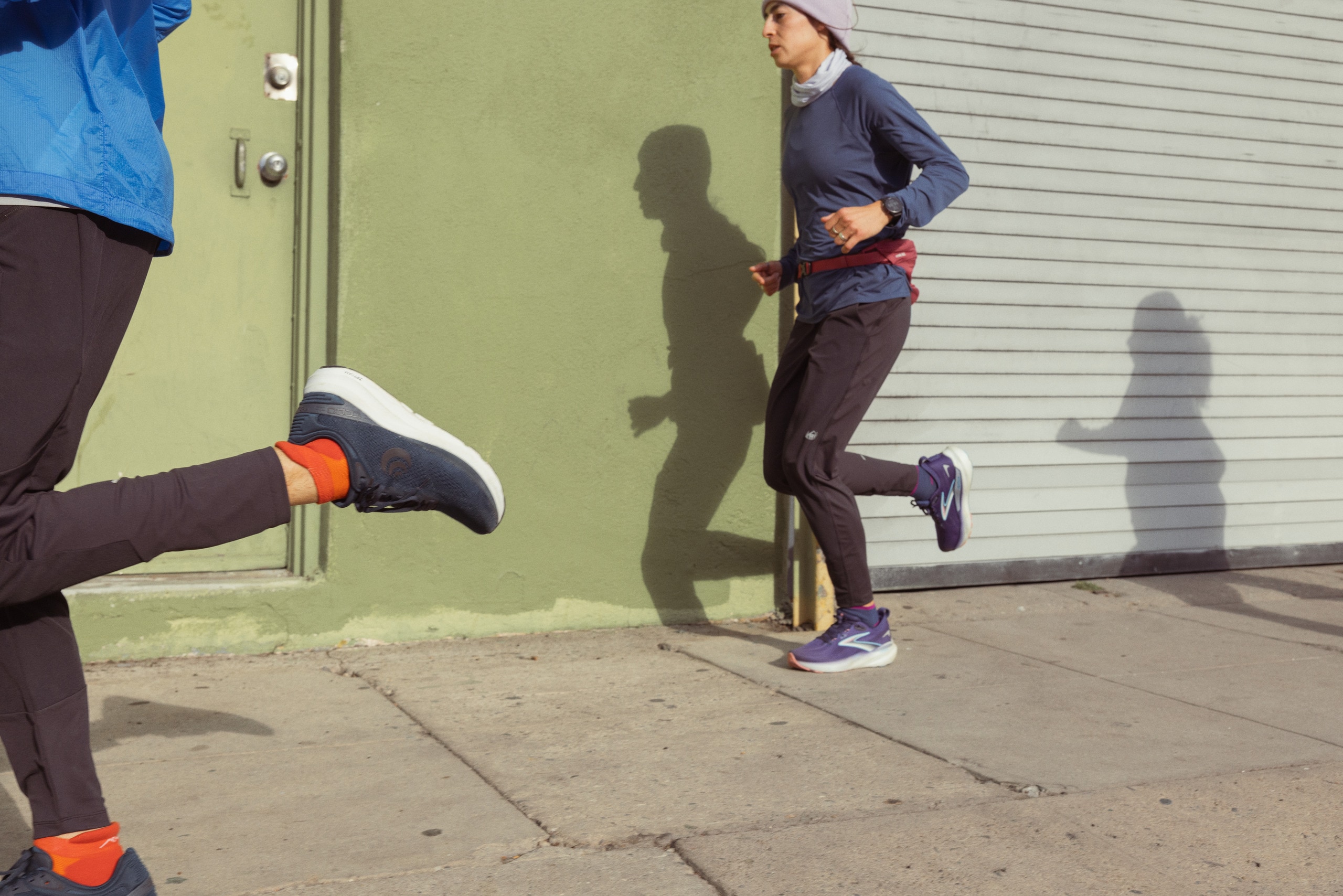 Three people are running along a sidewalk: One is passing by the camera and only their kick is visible, one is mid-stride, and one is seen in shadow.