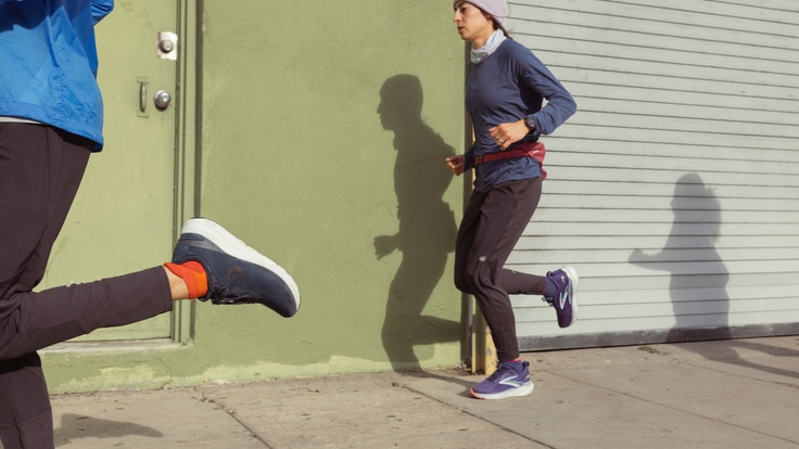 Three people are running along a sidewalk: One is passing by the camera and only their kick is visible, one is mid-stride, and one is seen in shadow.