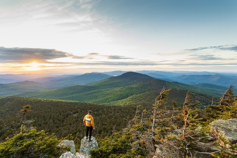 A hiker stands on a rock and overlooks a view of rolling green hills in the distance.