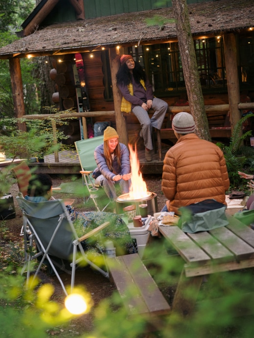 People gathered around a campfire near a rustic cabin in the woods, bundled in warm clothes.