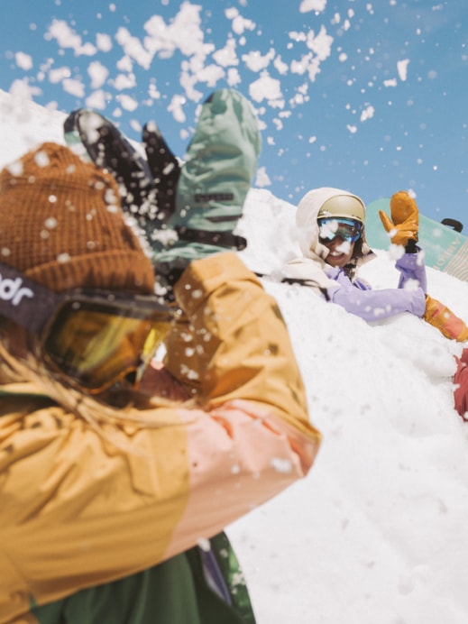 People in snow gear and goggles have a playful snowball fight on a sunny day.