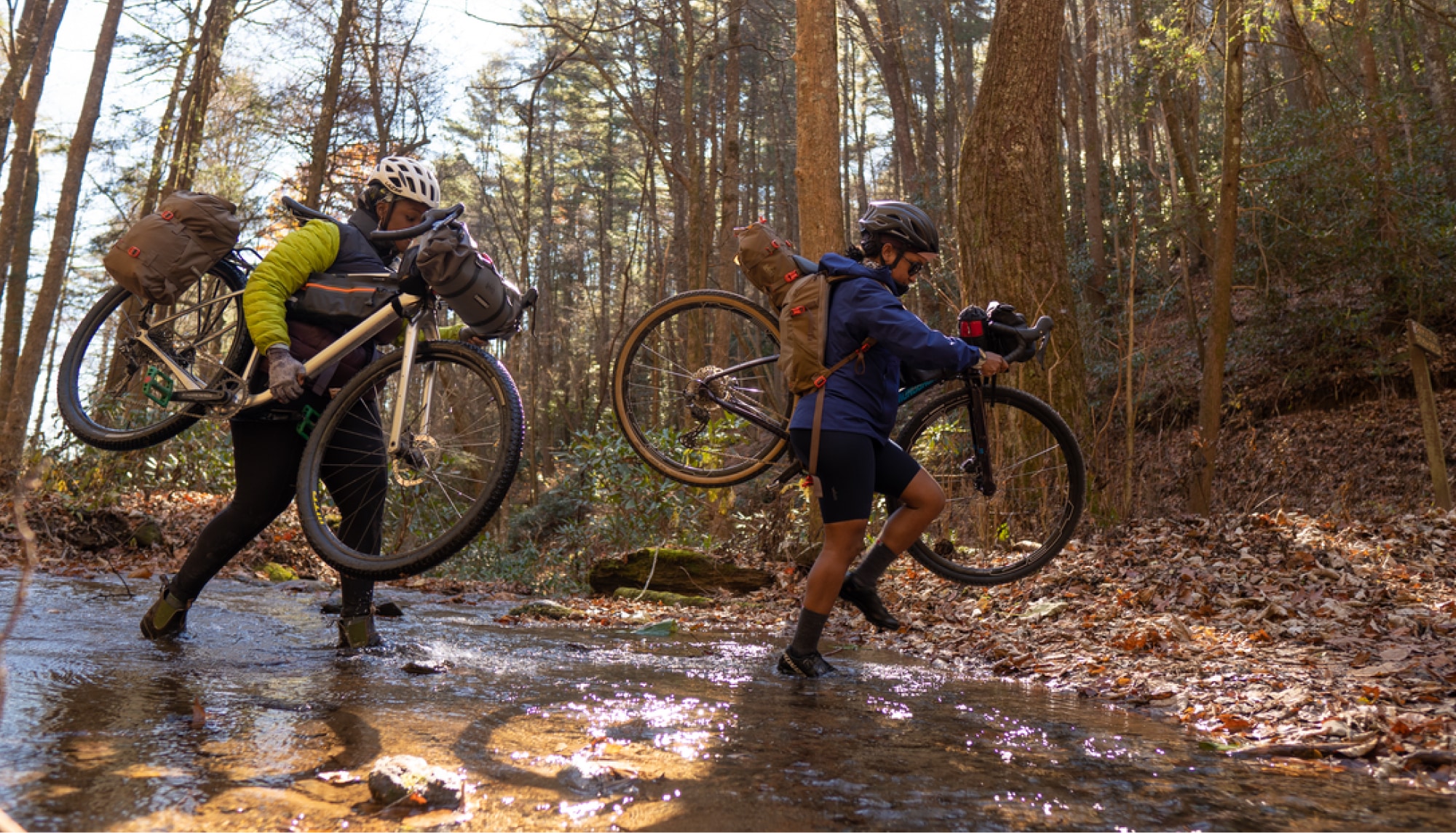 Two people carrying their bikes over a creek.