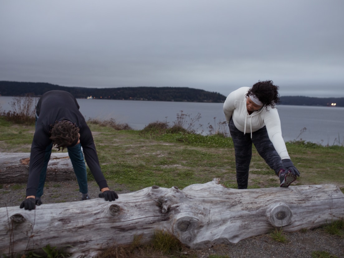 Two people stretching on a log near a body of water.