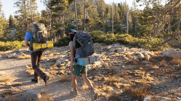 Two backpackers carrying Osprey backpacks hike a trail in the forest.