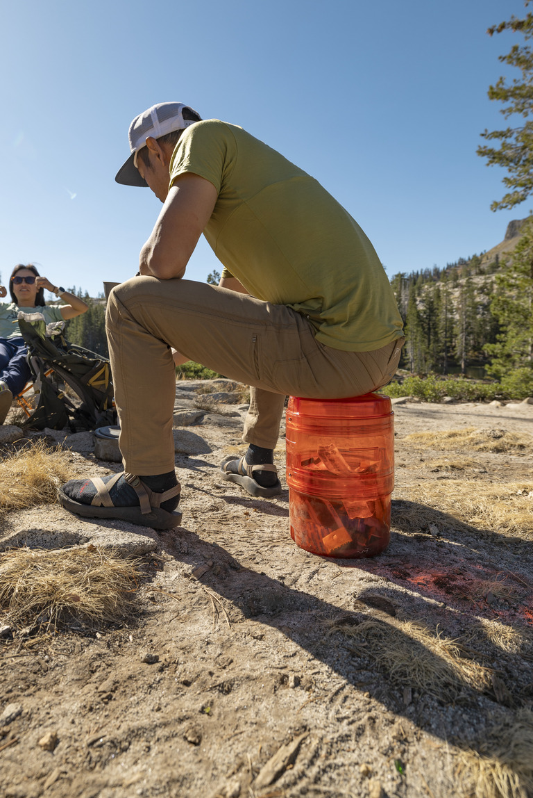 A backpacker sits on an orange bear-resistance food canister