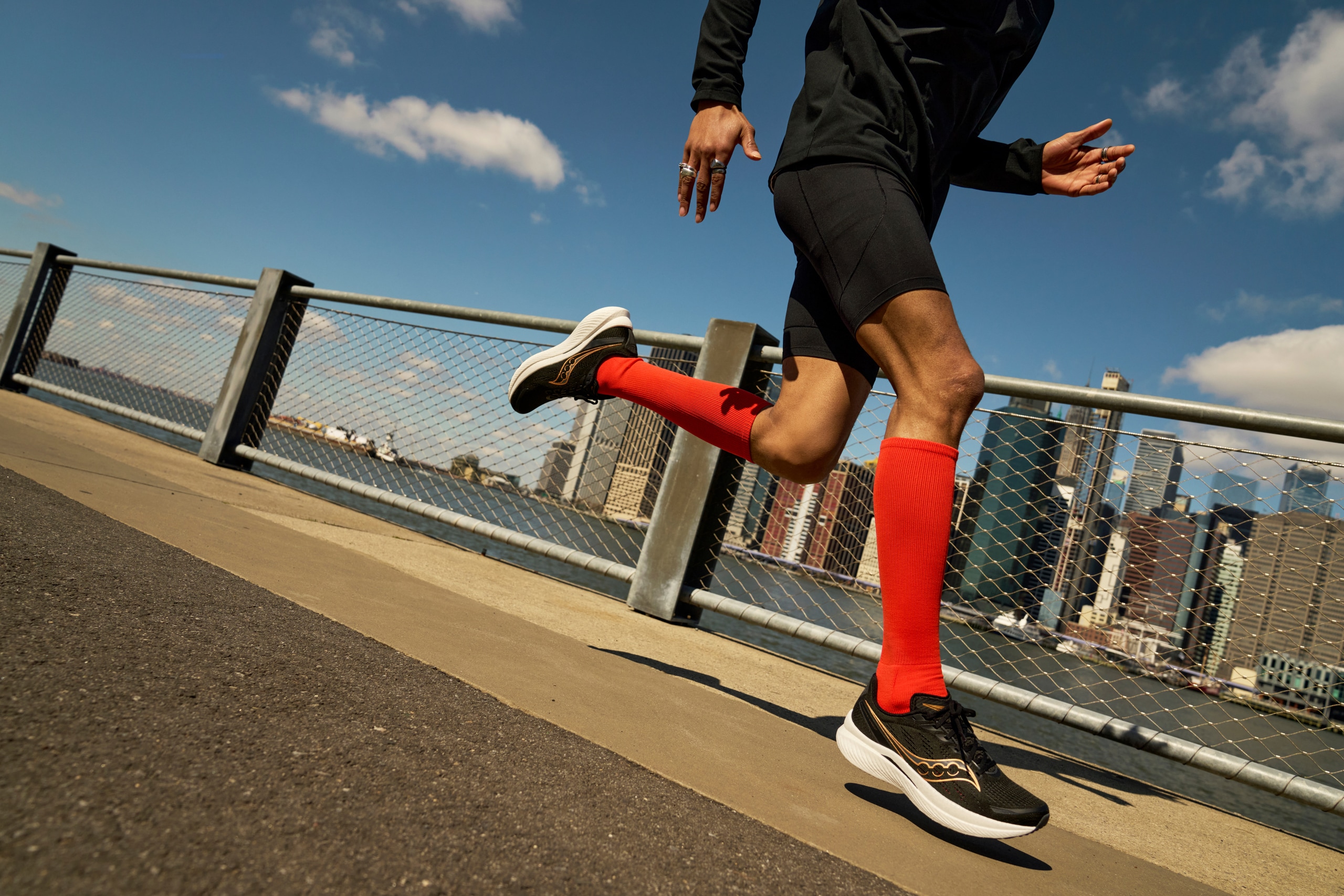 A runner in full flight across a city bridge, wearing black Saucony Endorphin Speed 3 road running shoes.