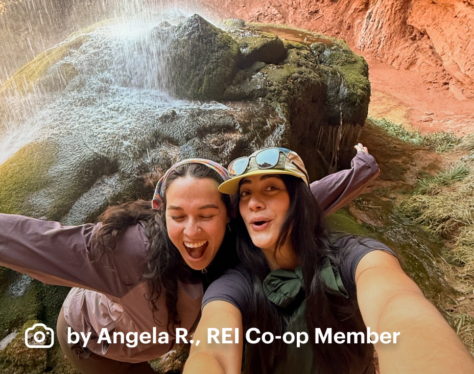 REI Co-op Member Angela R. and friend playfully pose under a canyon waterfall during a summer hike.