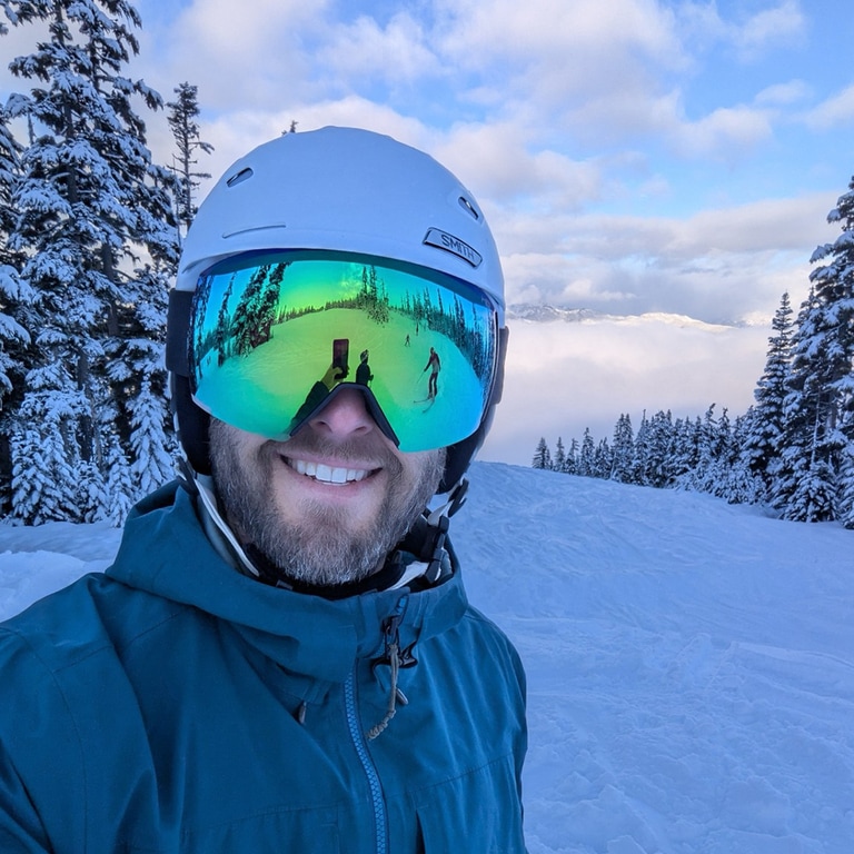 A skier wearing a helmet and goggles stands in front of a snowy landscape.