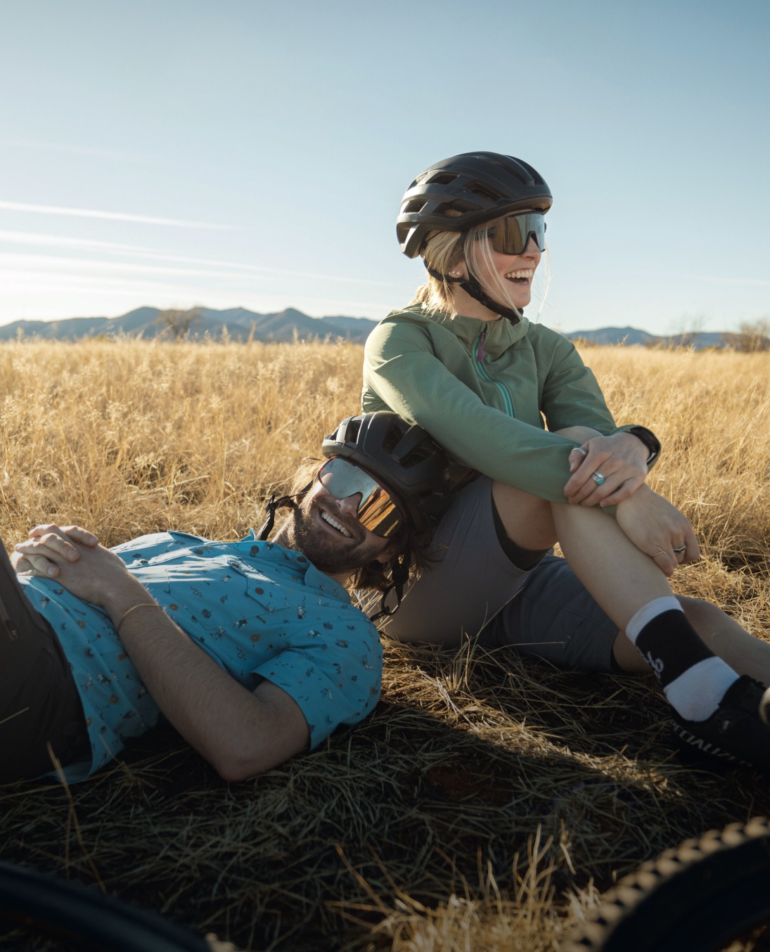 Two bikers stop to take a break and sit together in a field.