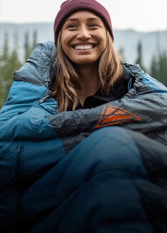 A camper in the Kelty Cosmic 20 sleeping bag sits on the ground and holds a mug