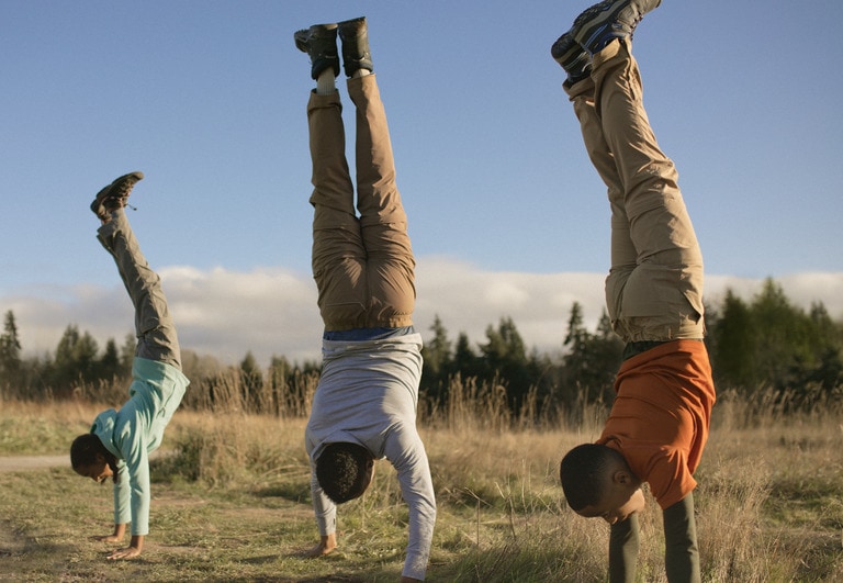 Three people in matching hiking pants do handstands in a field.