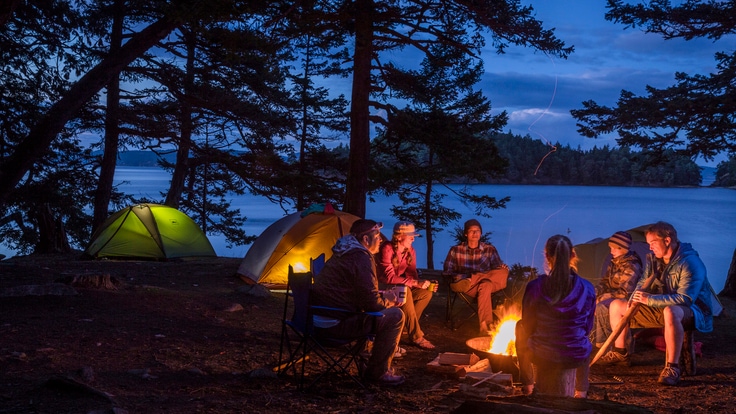 A group of people sit around a campfire at dusk, with their tents illuminated in the background