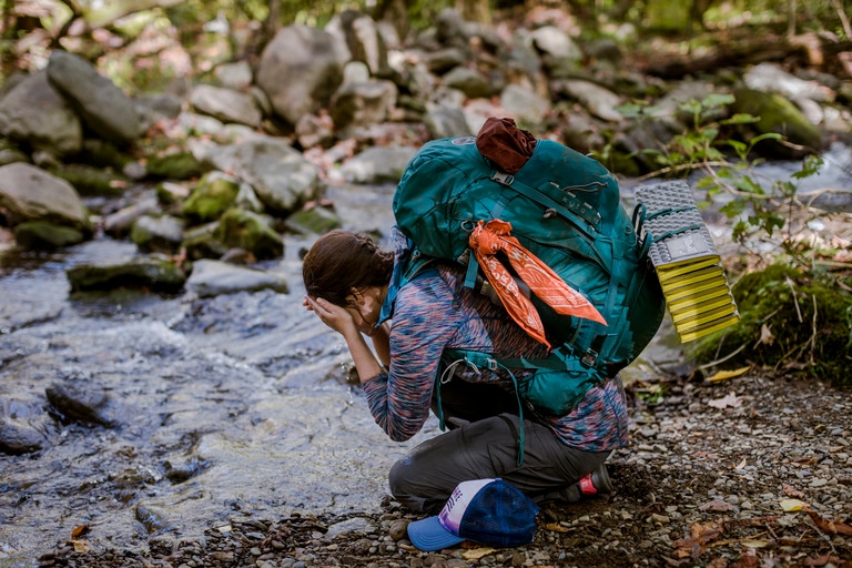  A person with a full backpacking pack kneeling over a stream and getting their face wet