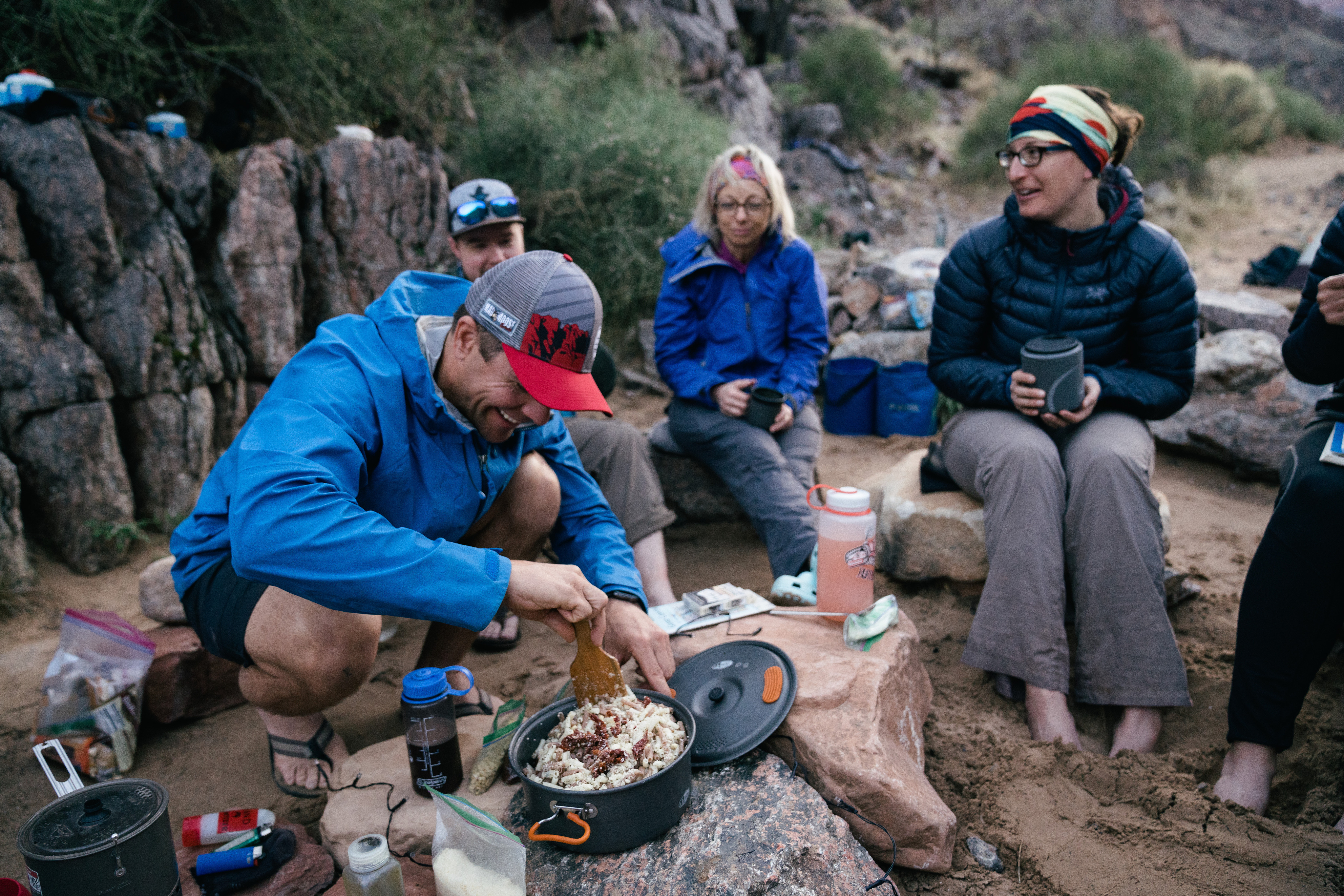 Five people sit on rocks with mugs as one person makes a backcountry meal.