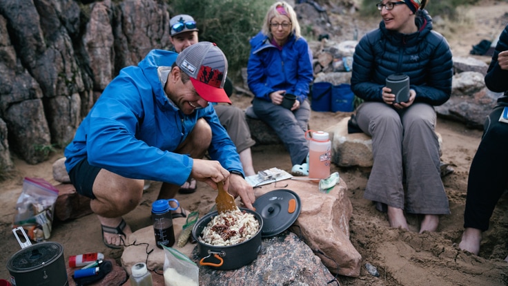 Five people sit on rocks with mugs as one person makes a backcountry meal.