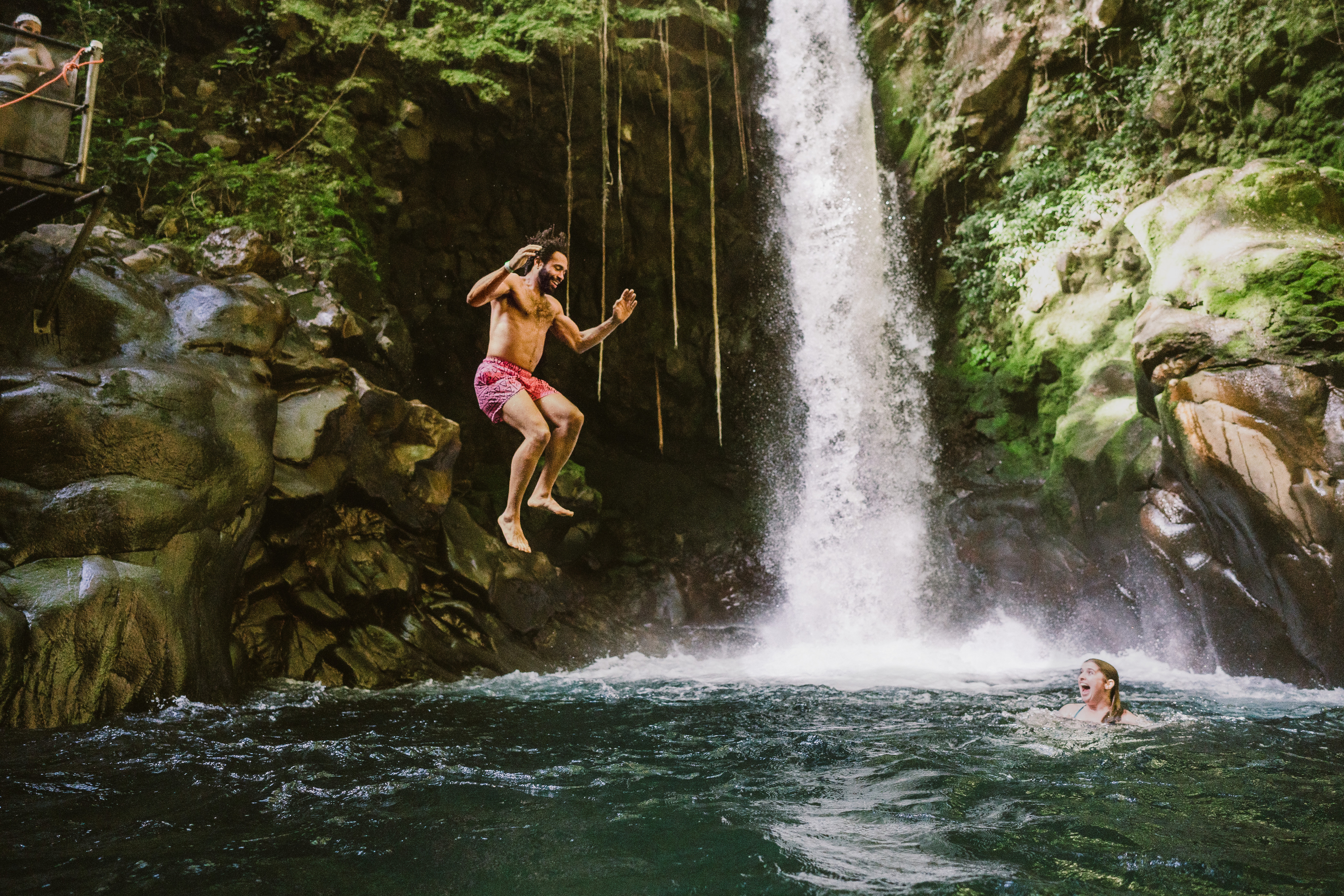 a man jumps off of a rock into a pool of water at the foot of a waterfall