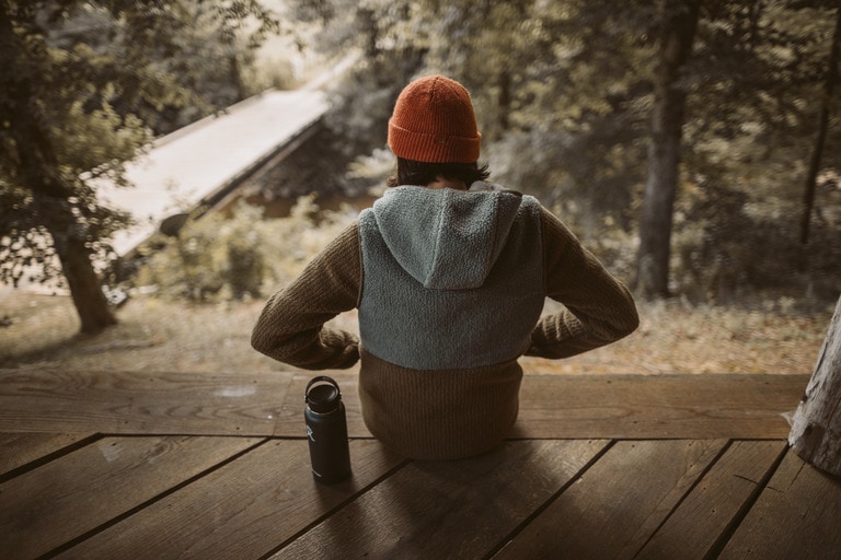 A person facing away from the camera, siting on a deck with a reusable water bottle, wearing a fleece hoodie and a knit watch cap.