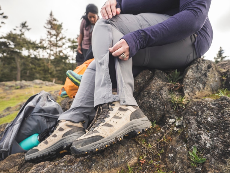 A person sits on a rock and unzips the leg of their hiking pants for ventilation.