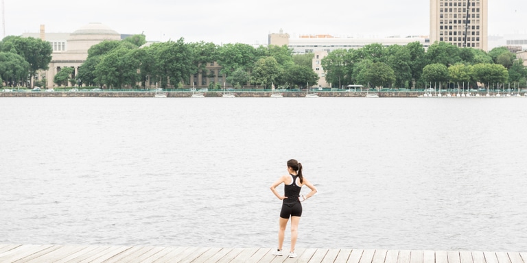 A woman in black running clothes stands on a pier with her hands on her hips, looking out to the water.